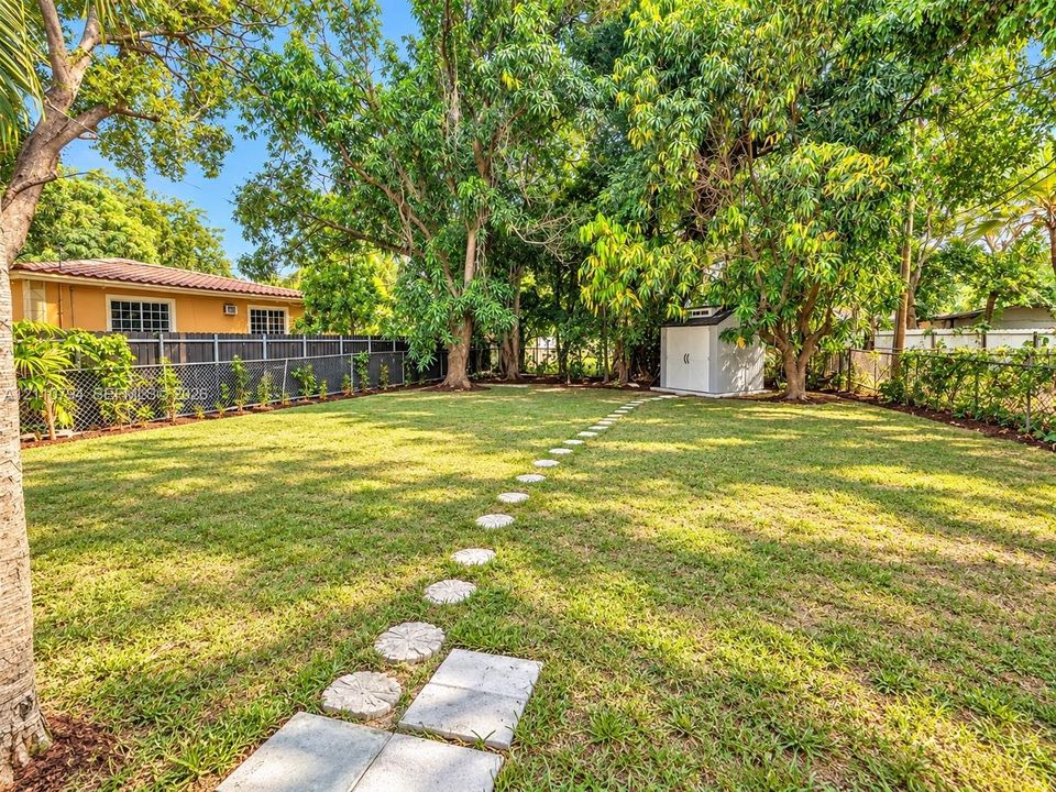 Expansive backyard featuring a grass lawn, mature trees, stone paver pathway, perimeter fencing, and a detached storage structure