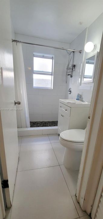Bathroom featuring large-format floor tiling, white subway wall tile, a glass-enclosed shower with pebble-stone flooring, and a white vanity with integrated basin