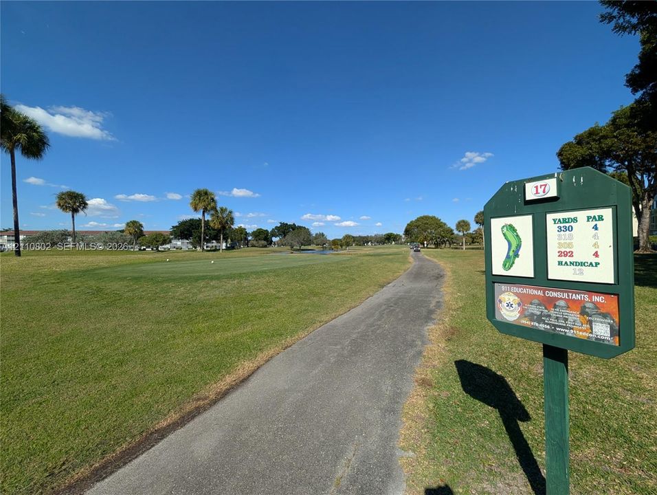 Paved cart path winding through a golf course featuring manicured turf, water feature, and mature palm trees