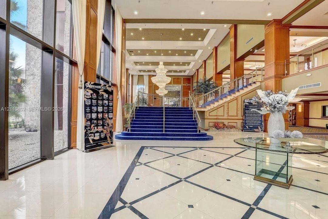 Grand foyer featuring expansive floor-to-ceiling windows, polished stone tile flooring with decorative inlay, a central multi-tier crystal chandelier, recessed coffered ceilings, and a staircase with an ornamental metal railing