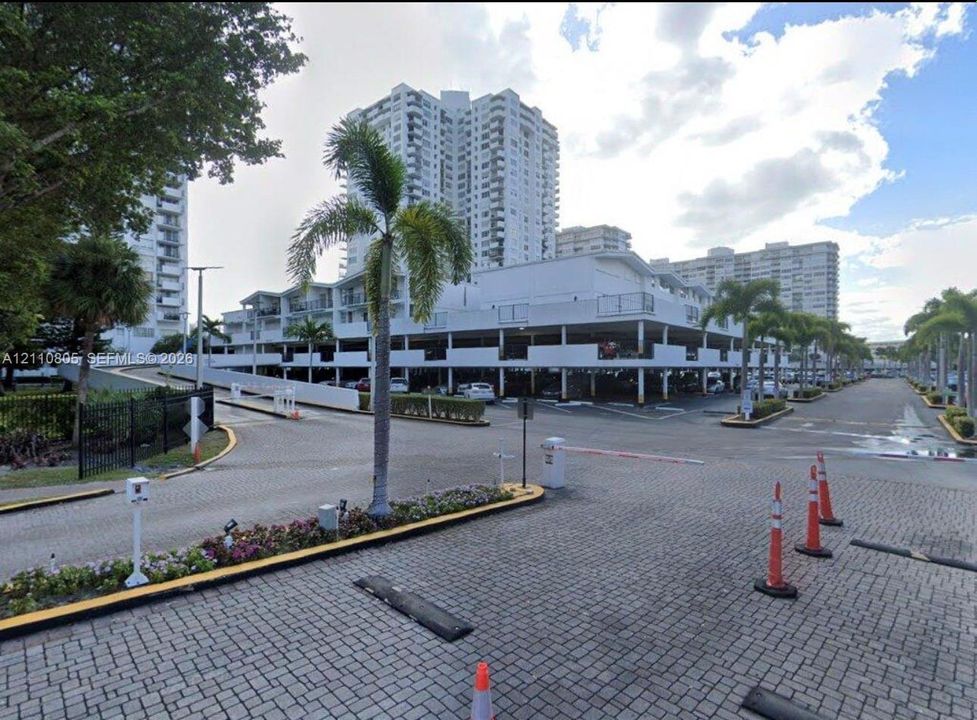 Covered parking structure featuring multiple levels, paved brick driveway, and manicured landscaping with palm trees