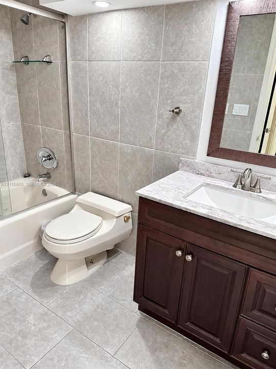 Bathroom featuring a dark wood-finish vanity with a stone-composite countertop, full-height tile wall surrounds, glass shower enclosure, and oversized ceramic floor tiling
