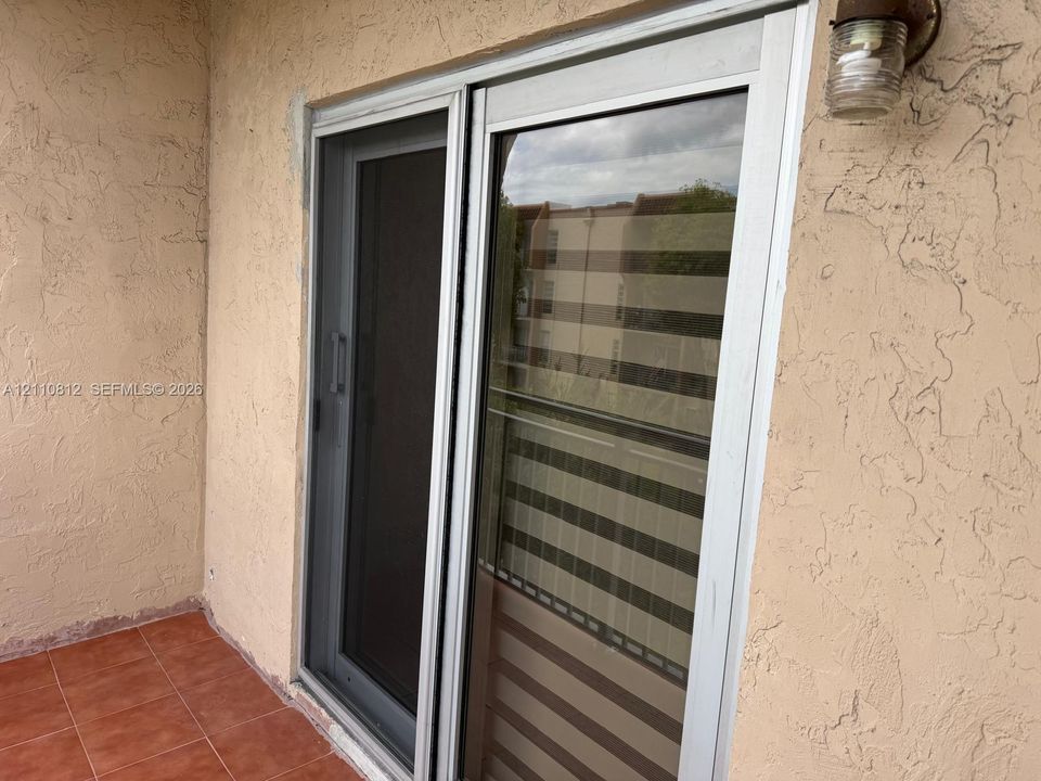 Sliding glass door with screen, stucco exterior wall, terra-cotta tile flooring, and wall-mounted exterior light fixture