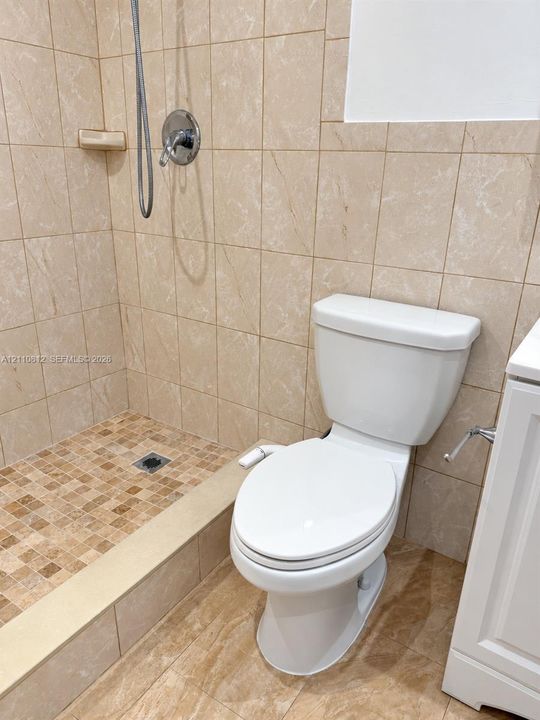 Walk-in shower featuring natural-tone wall tiles and small-scale mosaic floor tiling, complemented by a handheld shower head and a white porcelain toilet