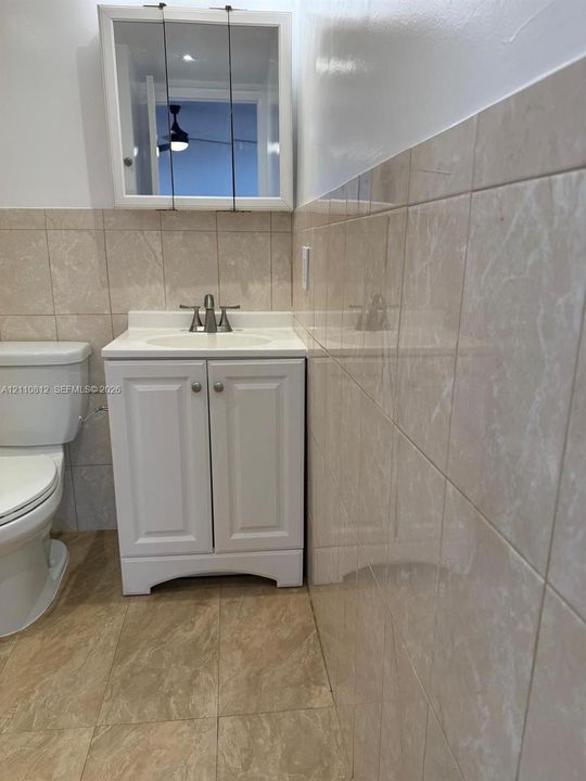 White vanity with integrated sink and two-handle faucet, wall-mounted mirrored medicine cabinet, and floor-to-ceiling tile surround