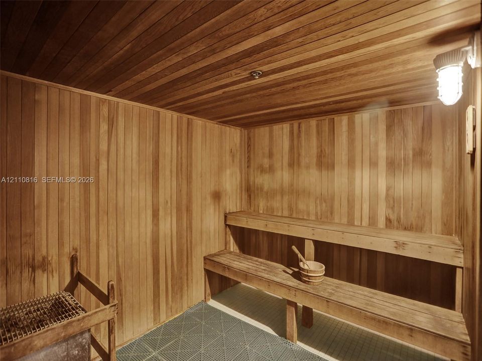 Sauna interior featuring natural wood-paneled walls and ceiling, multi-level tiered wooden benches, an integrated stone heater, and industrial-style wall-mounted lighting