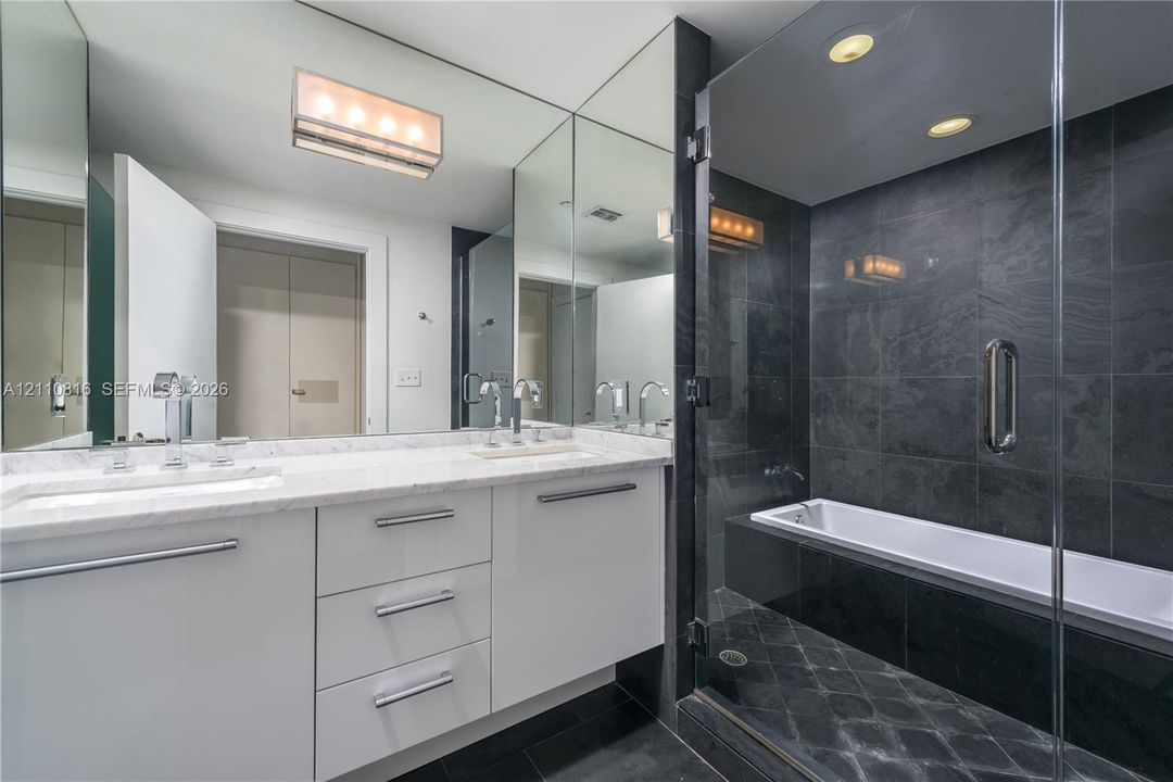 Bathroom featuring white cabinetry, stone-finish vanity countertop, dual undermount sinks with polished chrome fixtures, full-height vanity mirror, and a glass-enclosed bathtub with dark tile surround