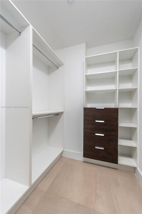 Walk-in closet featuring white built-in shelving, integrated hanging rods, a four-drawer dresser unit with dark-finish fronts, and light-toned flooring