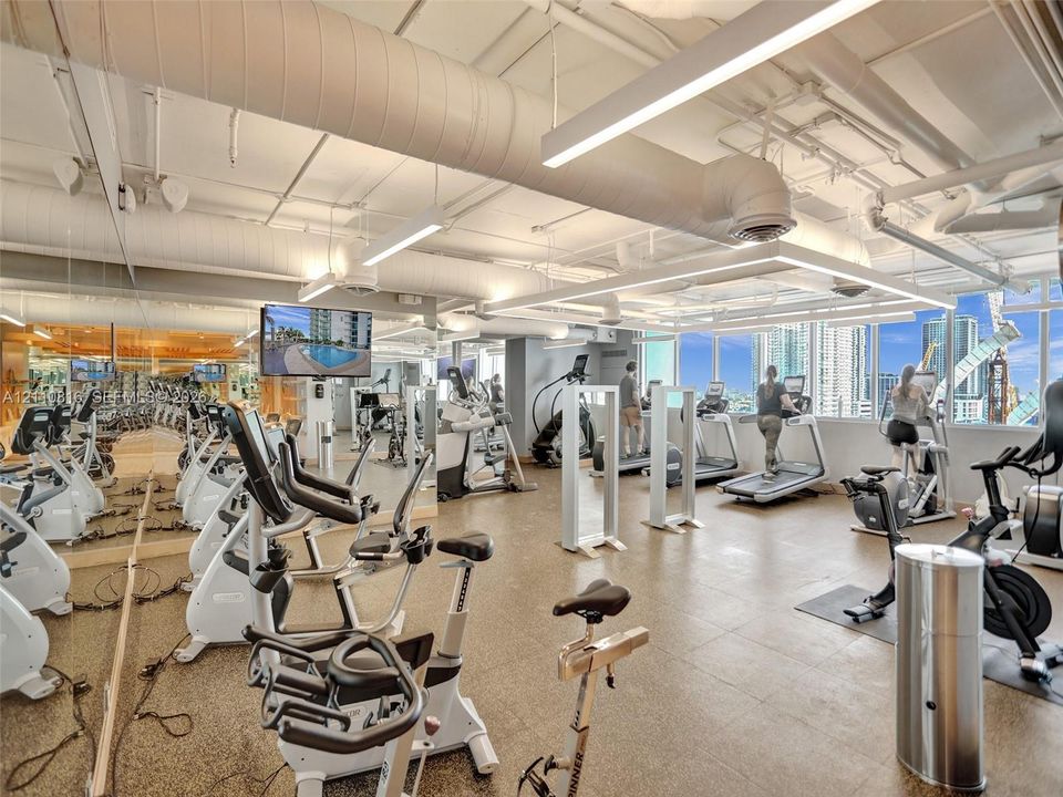 Fitness center featuring industrial-style exposed ductwork, wall-to-wall mirrored surfaces, expansive windows, linear overhead lighting, and stone-finish flooring