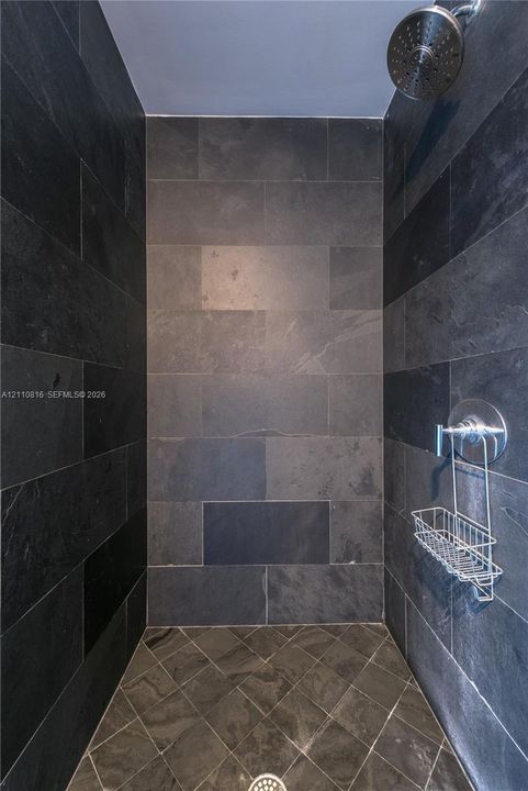 Walk-in shower enclosure featuring full-height charcoal stone tile walls, coordinating diamond-patterned tile flooring, a circular chrome showerhead, and a matching chrome pressure-balance valve