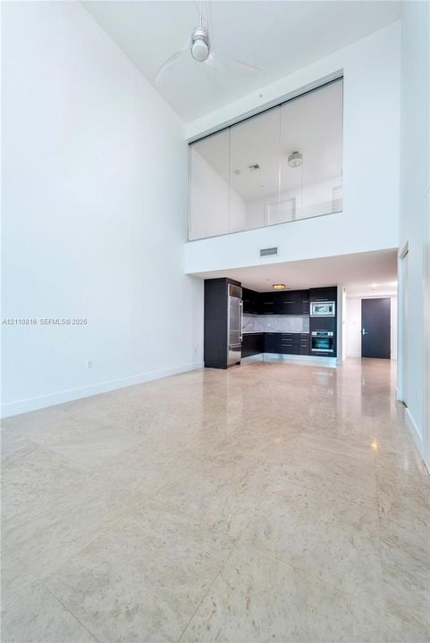 Double-height ceilings featuring an overhead fan, polished stone flooring, dark-toned cabinetry, stainless steel built-in appliances, and a glass-paneled interior wall