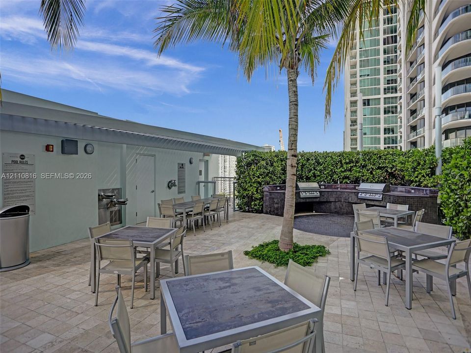 Outdoor terrace featuring stone-tile flooring, a built-in stone-finished kitchen station with integrated stainless steel grills, a manicured hedge perimeter, and palm landscaping