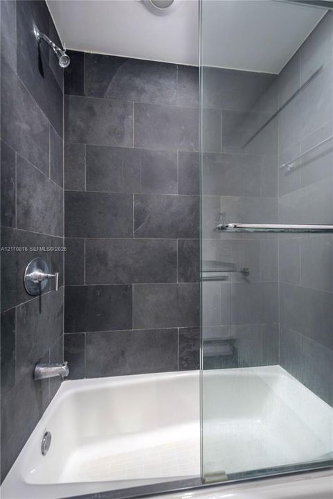 Bathtub with a glass enclosure, featuring large-format dark stone wall tile, chrome shower head, and wall-mounted chrome hardware