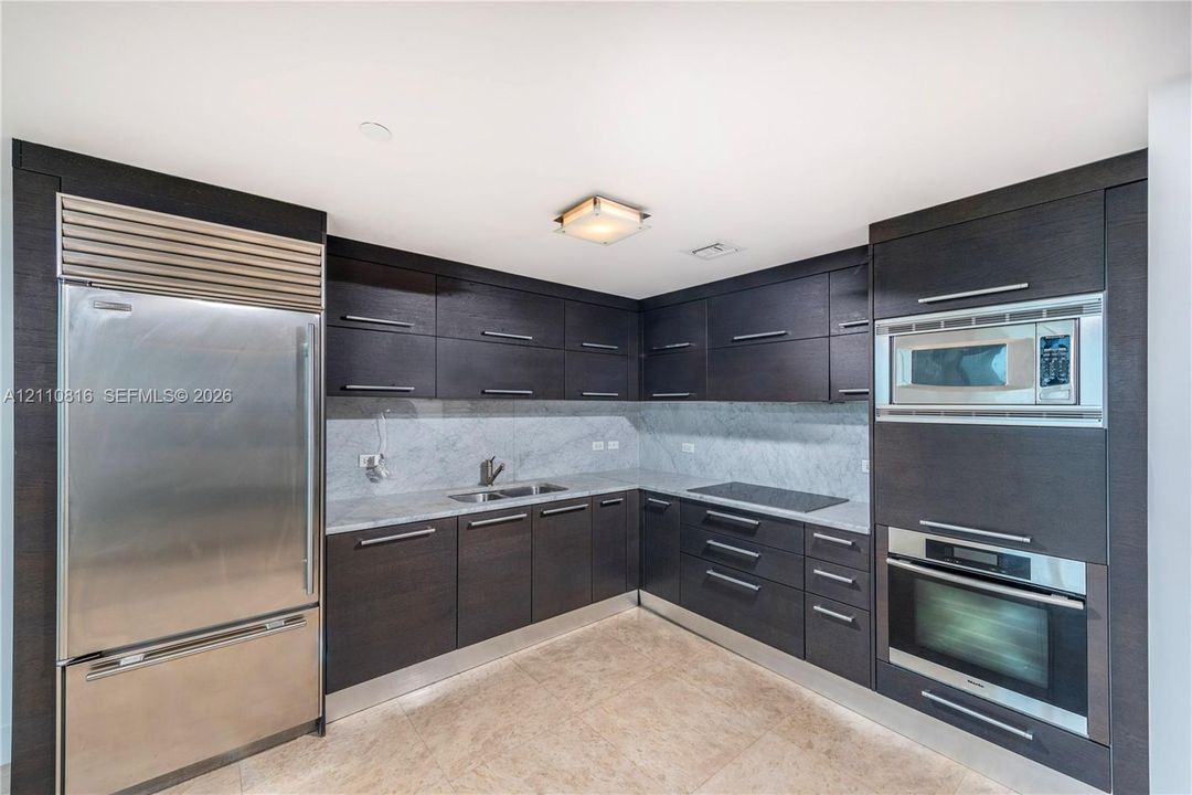 Kitchen featuring dark-toned flat-panel cabinetry, stone countertops, integrated stainless steel refrigerator, built-in microwave, and wall-mounted oven