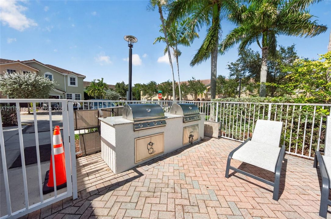 Outdoor kitchen featuring dual stainless steel grills, a stone-finish masonry surround, and a herringbone-pattern paver patio enclosed by metal fencing