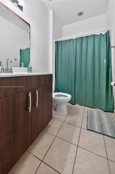 Bathroom featuring a dark wood-finish vanity with metal hardware, a wall-mounted mirror with an integrated light fixture, a toilet, tile flooring, and a bathtub enclosure with a rod