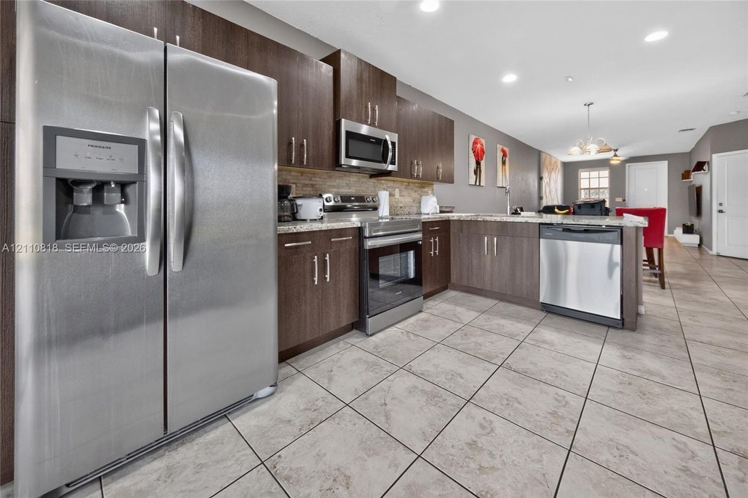 Kitchen featuring dark wood-finish cabinetry, stone-style countertops, tile flooring, and stainless steel appliances including a side-by-side refrigerator, microwave, and dishwasher