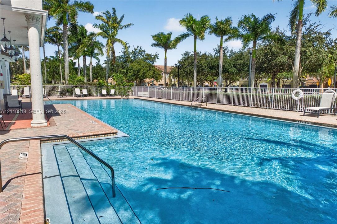 In-ground swimming pool featuring a brick-paver deck, built-in steps with a metal handrail, white aluminum perimeter fencing, and surrounding palm trees