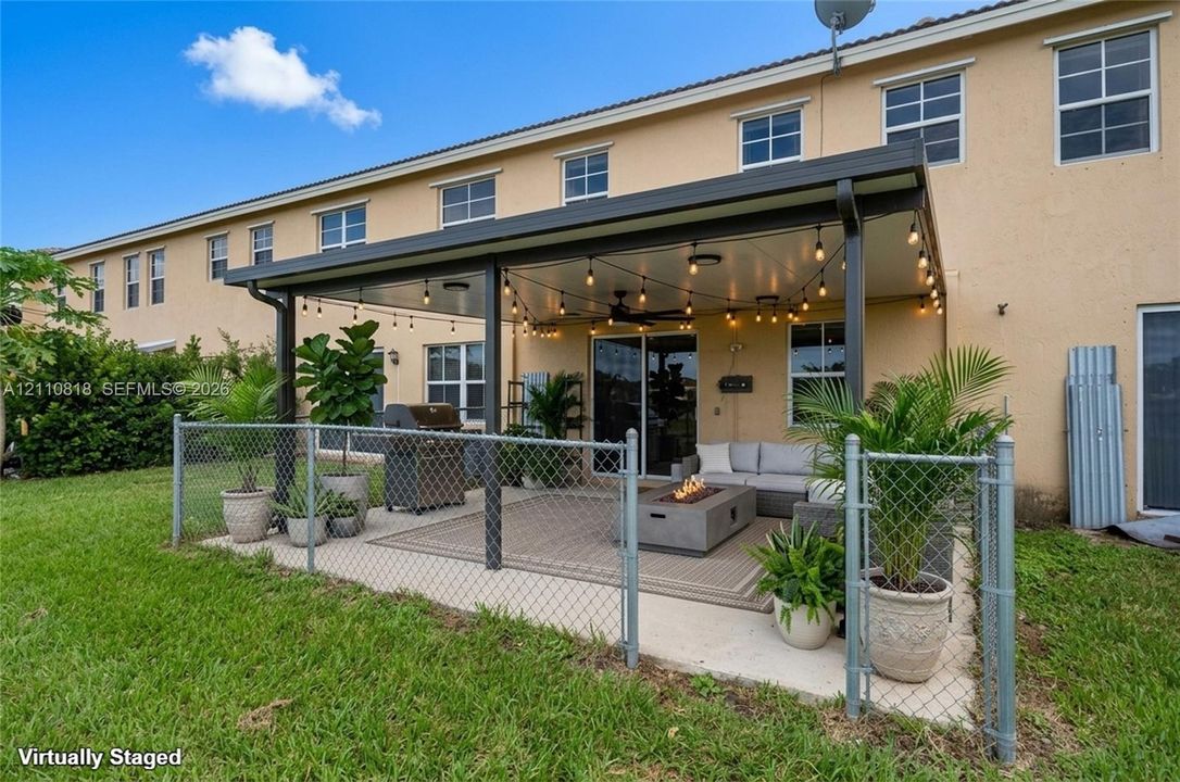 Covered concrete patio featuring a metal roof structure, ceiling fan, and string lighting, enclosed by chain-link fencing