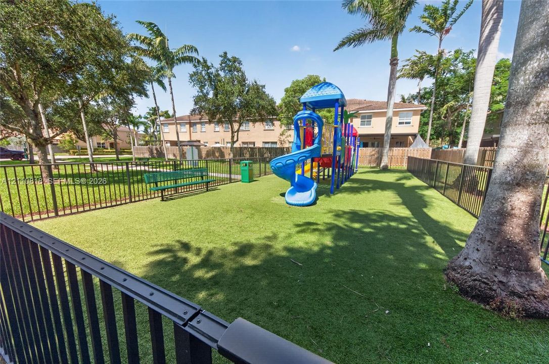 Fenced recreation area featuring artificial turf surfacing, a multi-component play structure, a metal bench, and mature palm trees