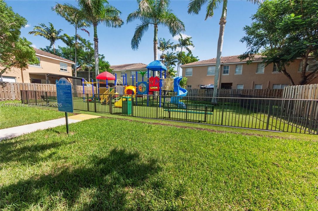 Fenced recreation area featuring a playground, manicured lawn, concrete walkway, and mature palm trees