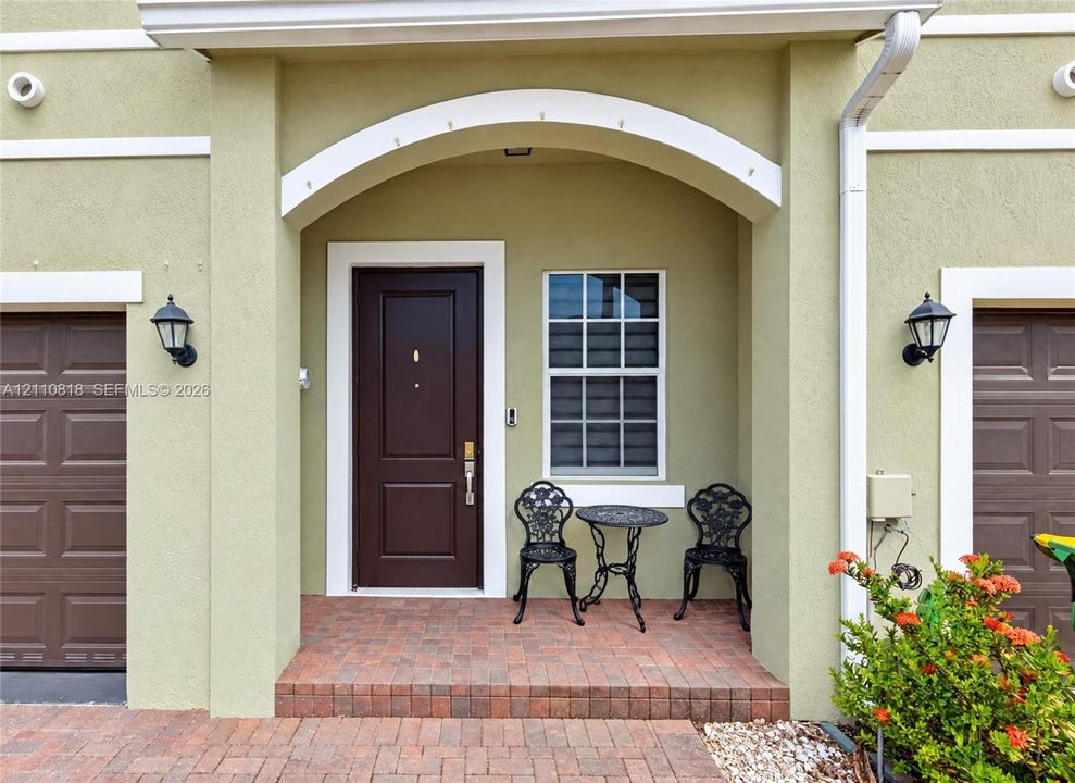 Stucco exterior entryway featuring a decorative arched portico, brick-paver flooring, solid-panel dark-finish entry door, and wall-mounted exterior lanterns