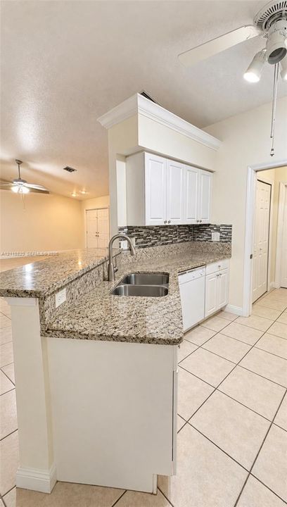 Kitchen featuring a peninsula with a stone-finish countertop, dual-basin stainless steel sink, tile backsplash, white cabinetry with crown molding, and ceramic tile flooring