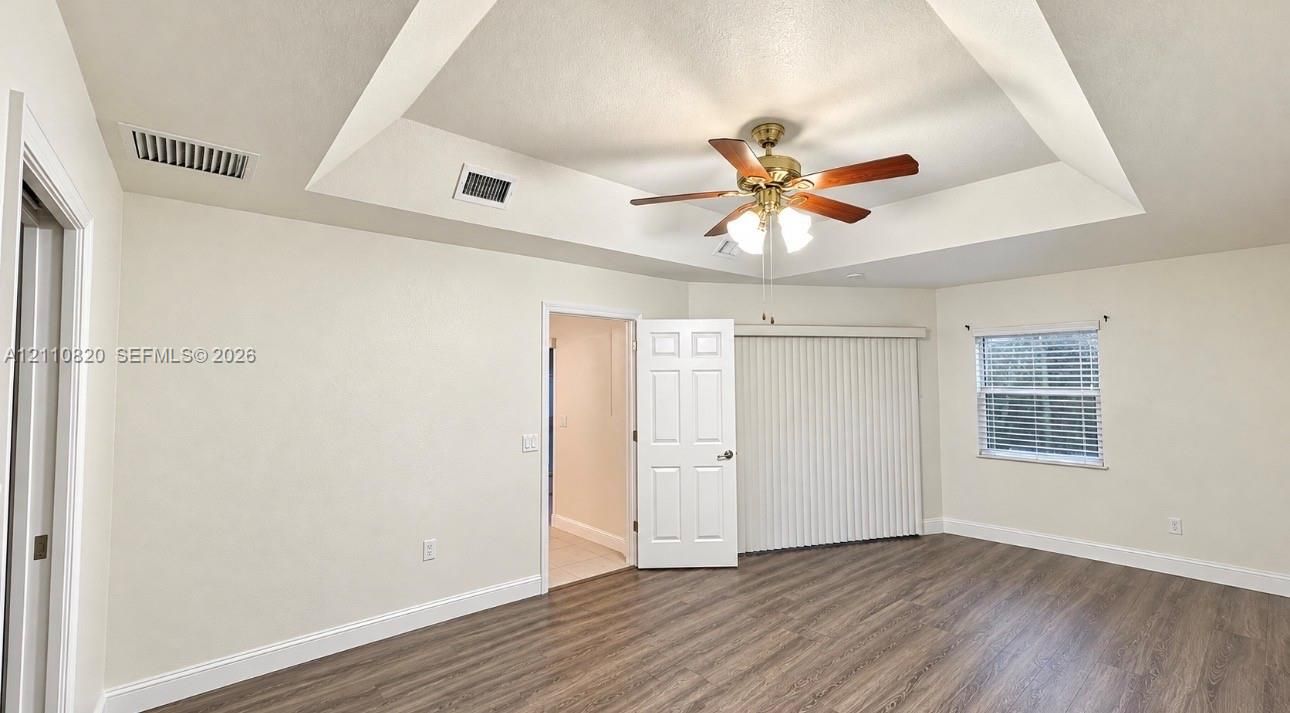 Tray ceiling featuring a central multi-blade ceiling fan with integrated lighting, wood-finish flooring, and vertical blinds