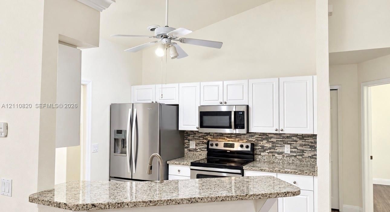 Kitchen featuring white shaker-style cabinetry, stone-finish countertops, stainless steel appliances, multi-toned mosaic tile backsplash, and a vaulted ceiling with a white ceiling fan