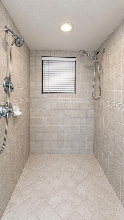 Walk-in shower featuring dual wall-mounted shower heads, ceramic tile surround with decorative inlay, recessed ceiling lighting, and window with integrated blinds