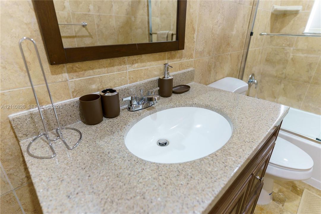 Vanity featuring an integrated oval sink, stone-composite countertop, dark wood-finish cabinetry, and neutral wall tile surround with a glass-enclosed shower