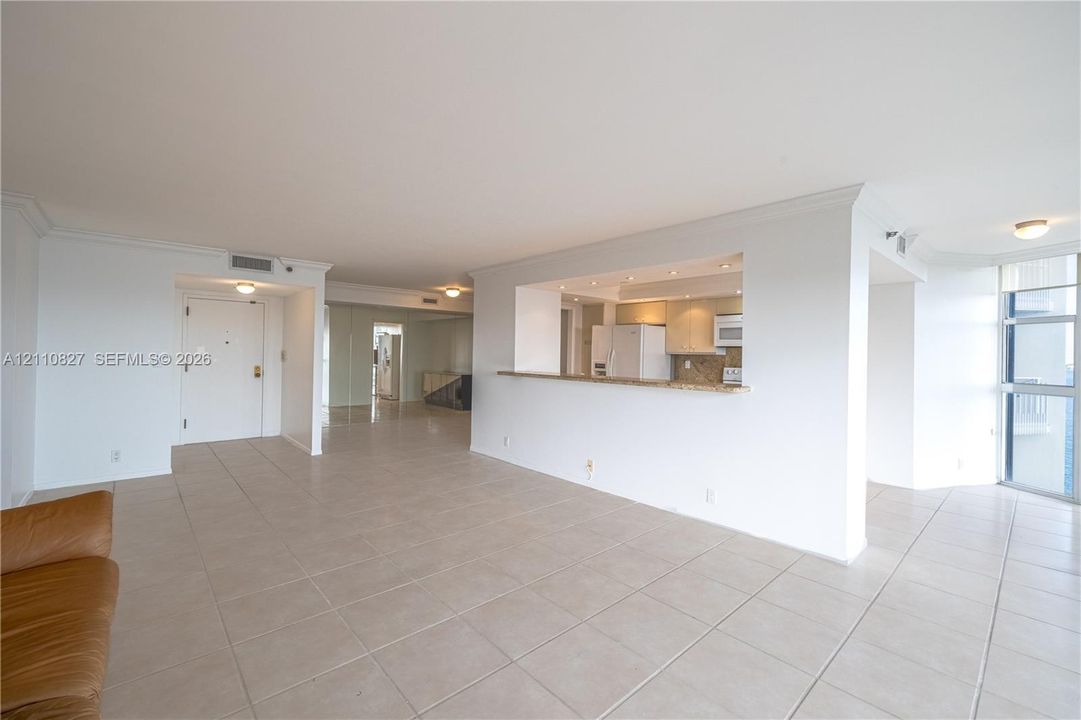 Open-concept living space featuring large-format tile flooring, crown molding, kitchen pass-through with stone countertop, and full-height windows