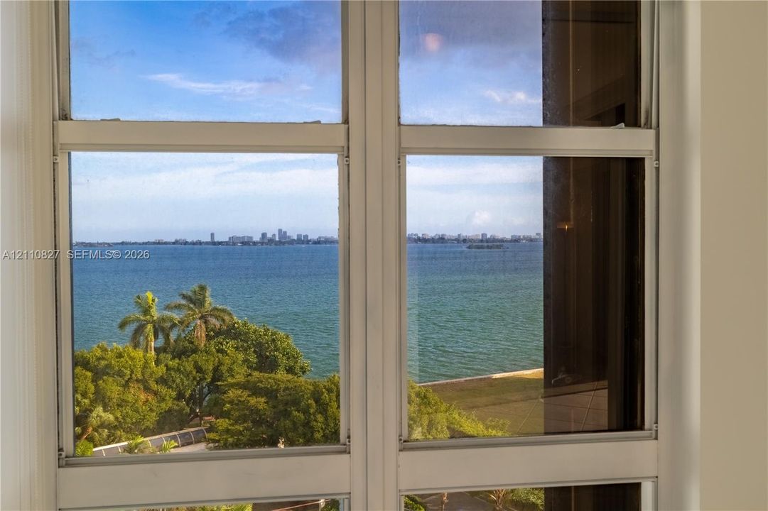 Double-pane window featuring a white frame, offering expansive water scenery, mature palm trees, and a distant cityscape horizon