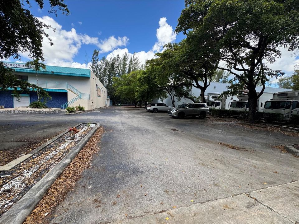 Expansive paved parking area featuring industrial structures, mature trees, and concrete curbing