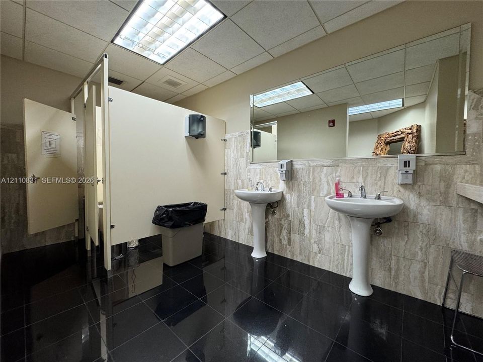 Restroom featuring two white pedestal sinks, full-width wall mirror, stone-tile wainscoting, and polished dark-tone floor tile
