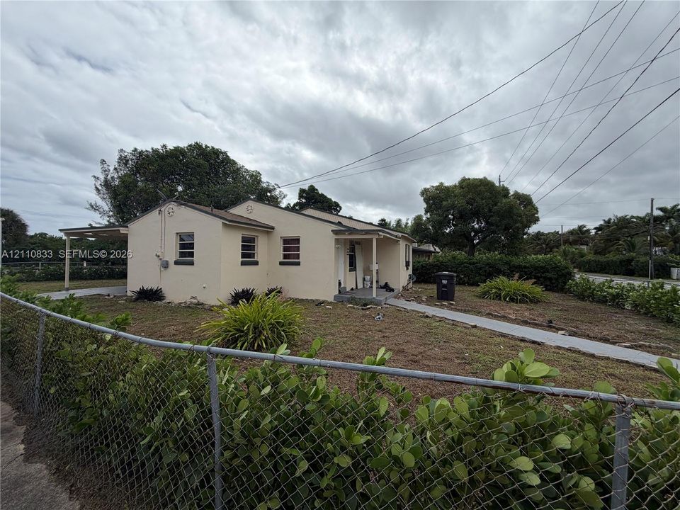 Single-story residence featuring a light-colored stucco exterior, attached carport, concrete walkway, and chain-link perimeter fencing