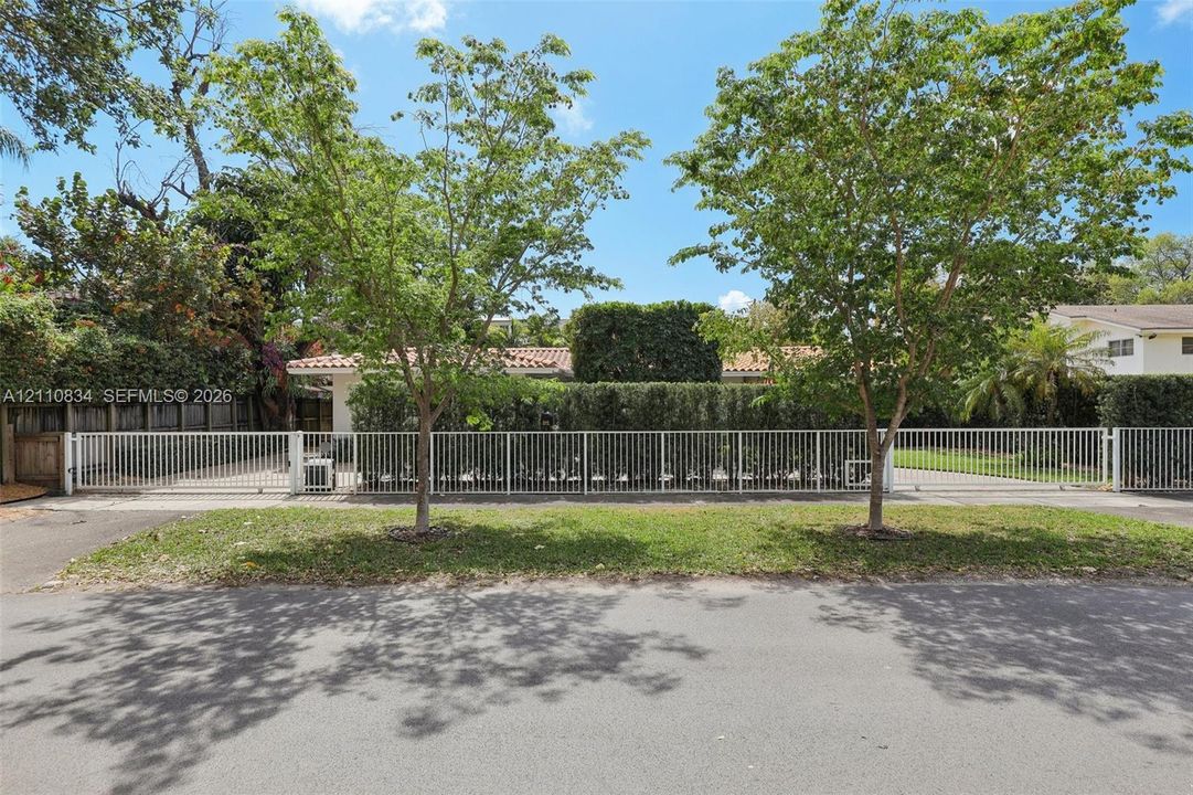 White metal perimeter fencing, gated driveway access, mature landscaping, and established trees