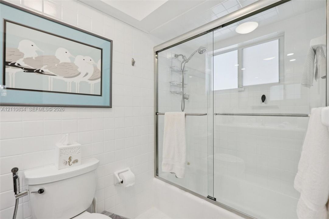 Bathroom featuring white tile wall surround, glass-enclosed shower-tub combination, chrome plumbing fixtures, and a recessed ceiling light