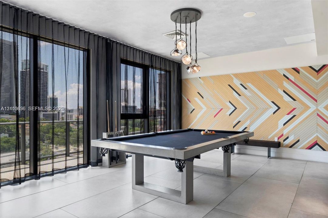 Modern recreation room featuring a regulation pool table, geometric wood-finish accent wall with multi-colored chevron detailing, floor-to-ceiling glass doors with dark framing, polished concrete flooring, and a contemporary pendant light fixture suspended from the ceiling