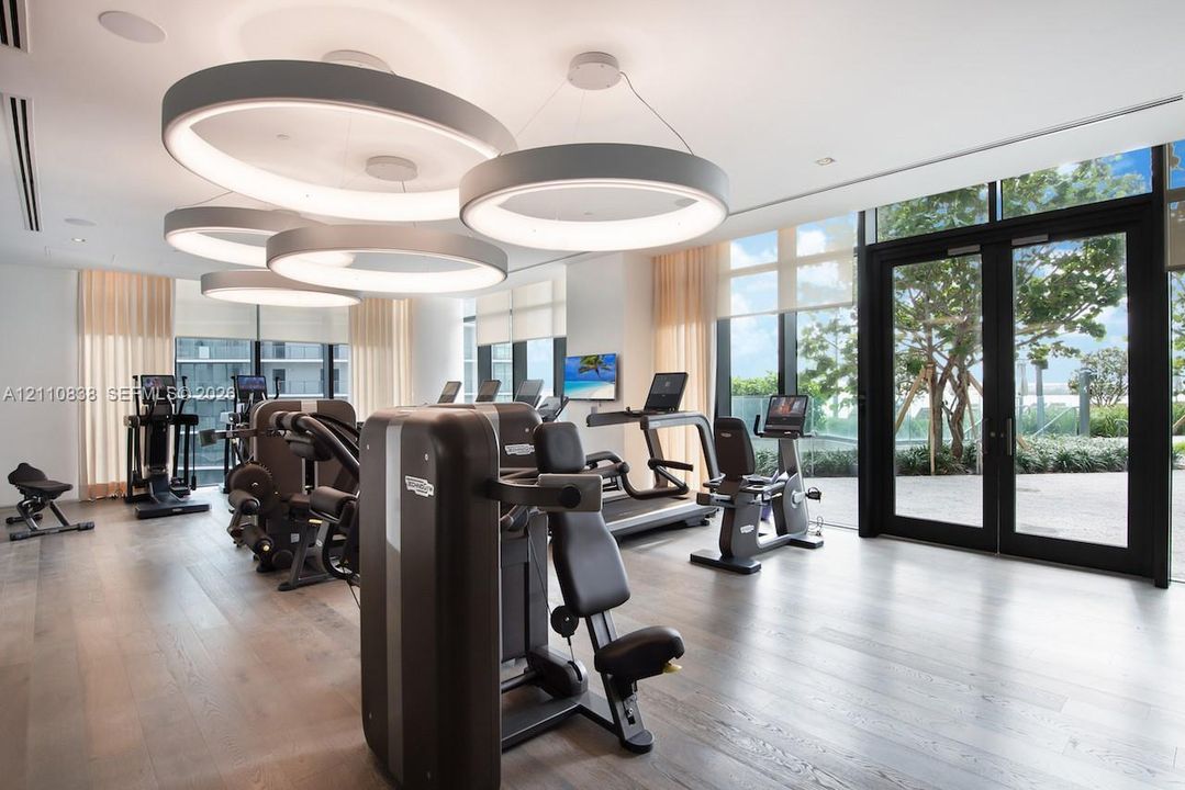 Fitness center featuring wood-finish flooring, floor-to-ceiling windows, circular LED pendant lighting, and double-door exterior access