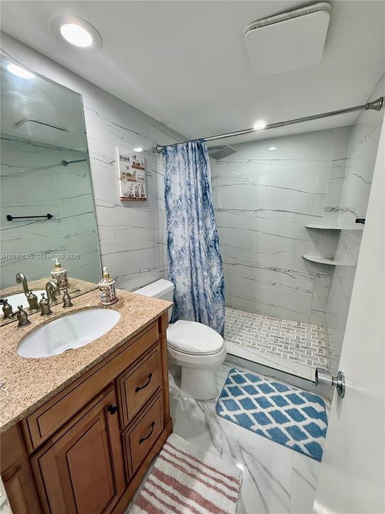 Bathroom featuring a wood-finish vanity with a stone-composite countertop, undermount sink, recessed lighting, and a walk-in shower with stone-patterned wall tile and built-in shelving