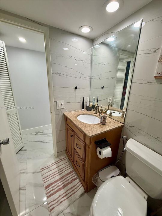 Full-height wall tiling featuring stone-patterned surfacing, vanity with integrated stone countertop and oval undermount sink, wall-spanning vanity mirror, recessed ceiling lighting, and large-format floor tiling