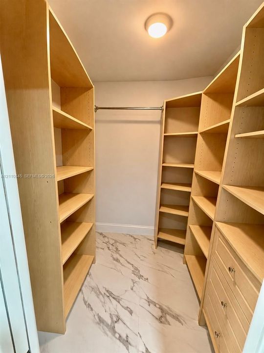 Walk-in closet featuring light-toned custom shelving, integrated drawer stack, metal hanging rod, and large-format tile flooring with a marble-patterned finish