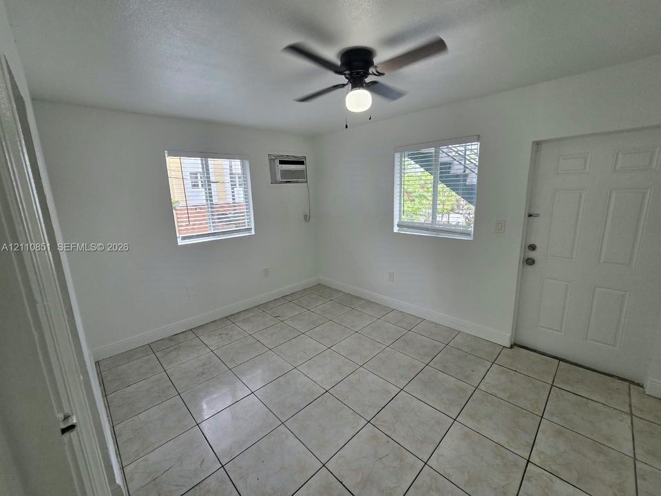 Neutral-toned tile flooring, white wall finishes, two windows with horizontal blinds, built-in wall-mounted air conditioning unit, and a ceiling fan with integrated light fixture