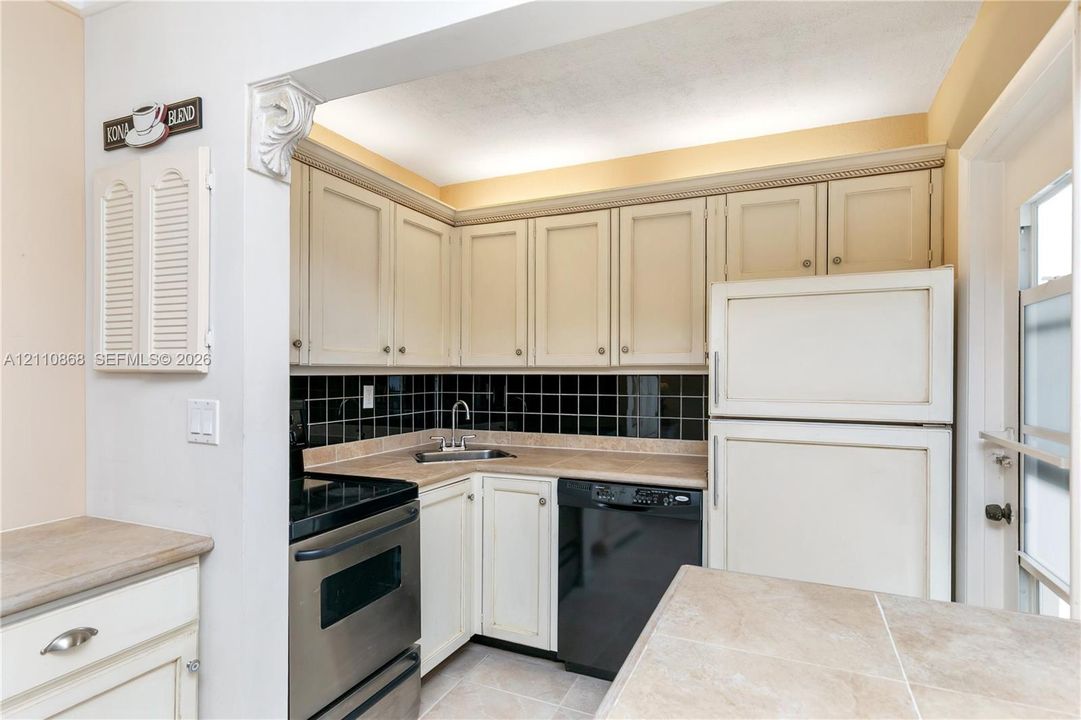 Kitchen featuring light-toned shaker cabinetry, black tiled backsplash, stone-finish countertops, and integrated stainless steel and black appliances