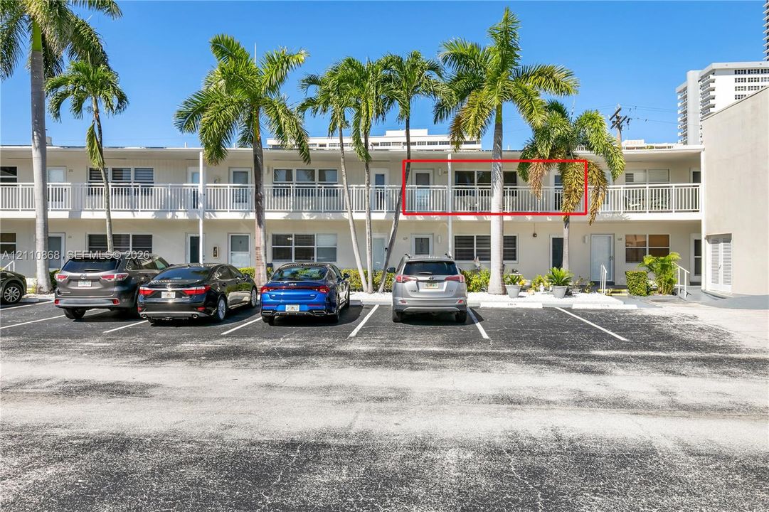 Two-story residential building featuring a second-level exterior balcony with white metal railings, integrated surface parking, and mature palm trees