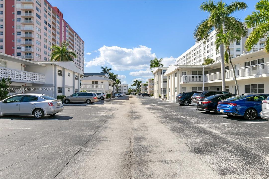 Asphalt parking area featuring multi-story residential buildings, white exterior facades, horizontal balcony railings, and palm trees