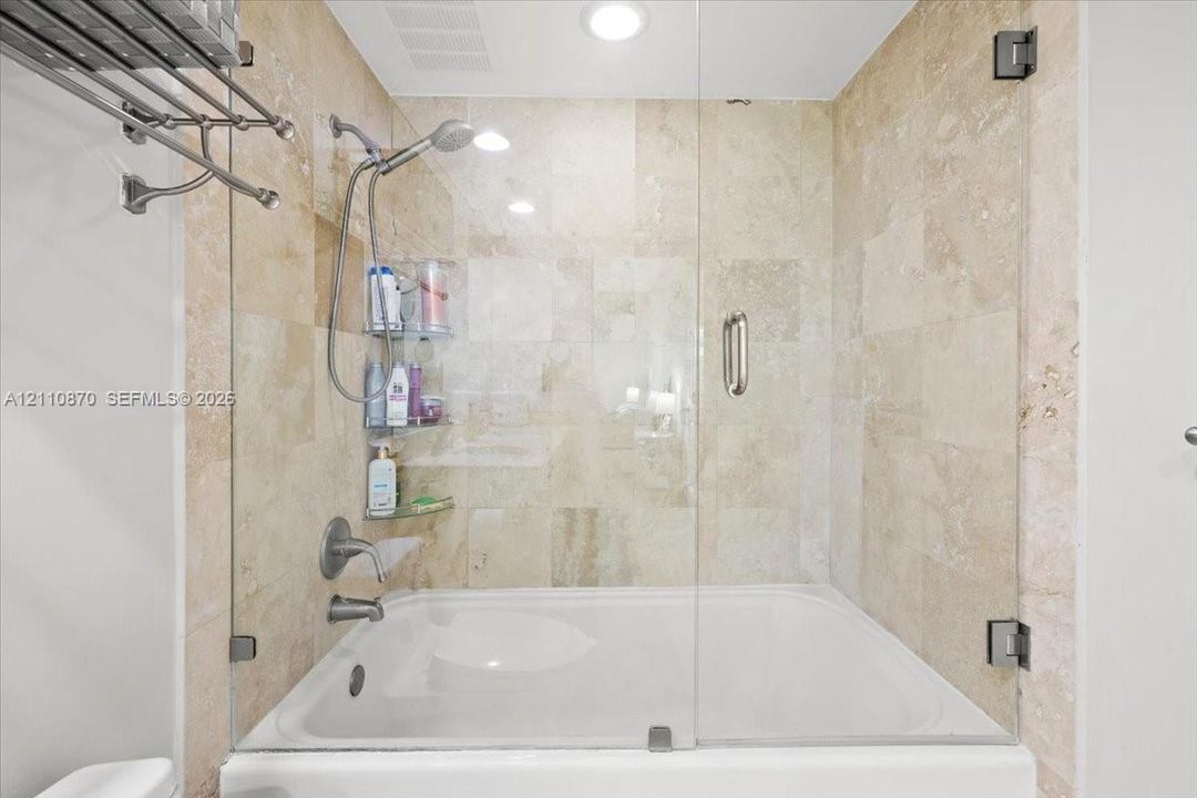 Bathtub with a glass enclosure, wall-mounted shower head, recessed lighting, and tiled wall surround
