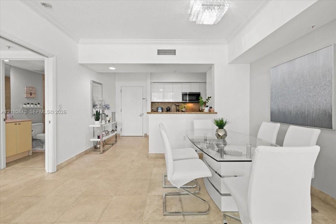 Open-concept floor plan featuring neutral stone-finish tile flooring, a tiered tray ceiling with a crystal-style chandelier, and high-gloss white cabinetry