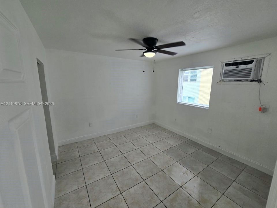 Neutral-toned tile flooring, white wall finishes, black ceiling fan with integrated lighting, and a wall-mounted air conditioning unit